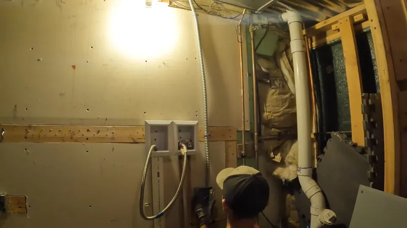 Worker crouching in a basement beside a white PVC drain stack and framed walls