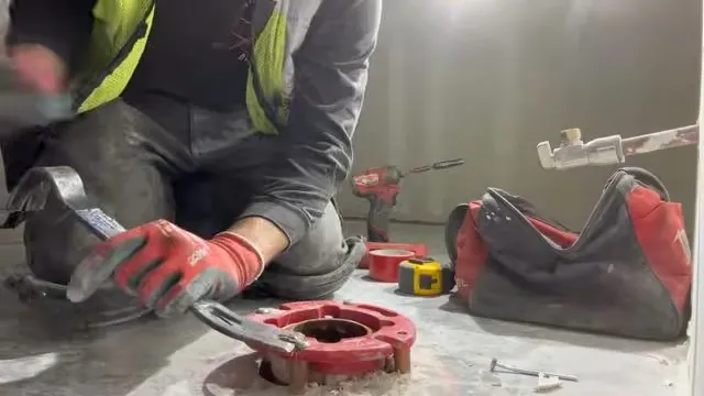 Worker in a hi-vis shirt kneeling at a red toilet flange on a concrete floor with a tool bag and drill